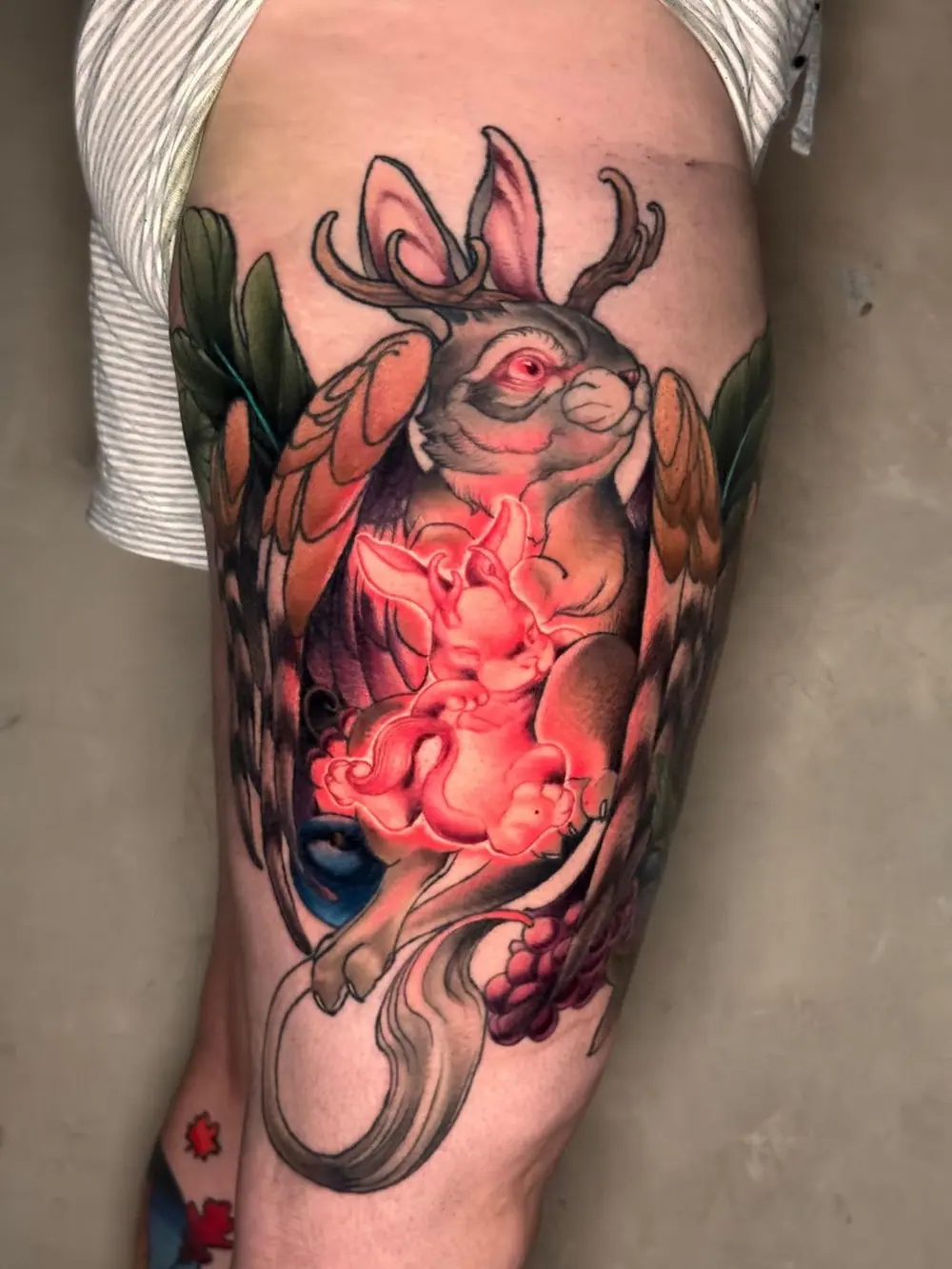 Tatuaje en el muslo: un jackalope, liebre con cuernos, que abraza un conejito rojo y blanco que brilla
